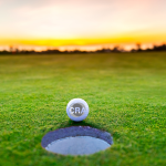 A golf ball with the CRA logo sits on the edge of a hole on the green. The sun is setting in the background.